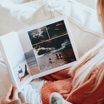 A woman sitting comfortably and browsing a luxury layflat photo book with a linen fabric cover, displaying travel memories from a road trip