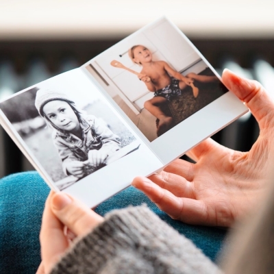 Hands holding an open Polaroid-style photo book showing retro prints of children — one in black and white outdoors, and one playing indoors with a wooden spoon.