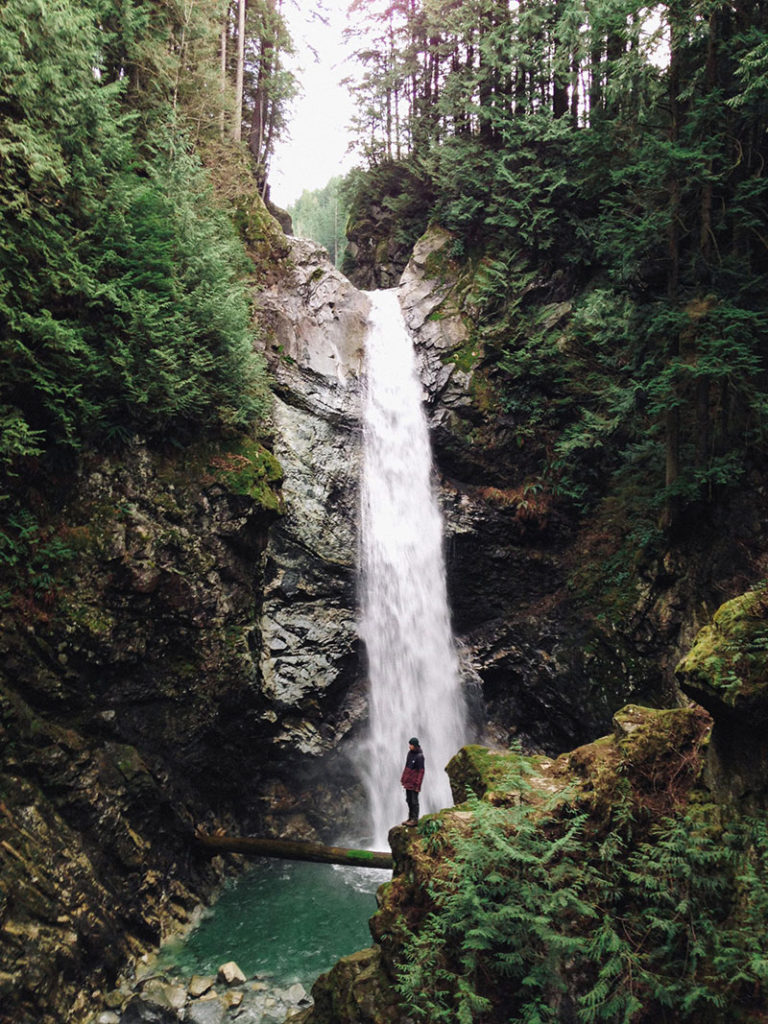Photo shows a woman in a red coat stood in front of a waterfall. The waterfall acts as a frame and shows how effective the iphone photography tip Frame Your Subject is