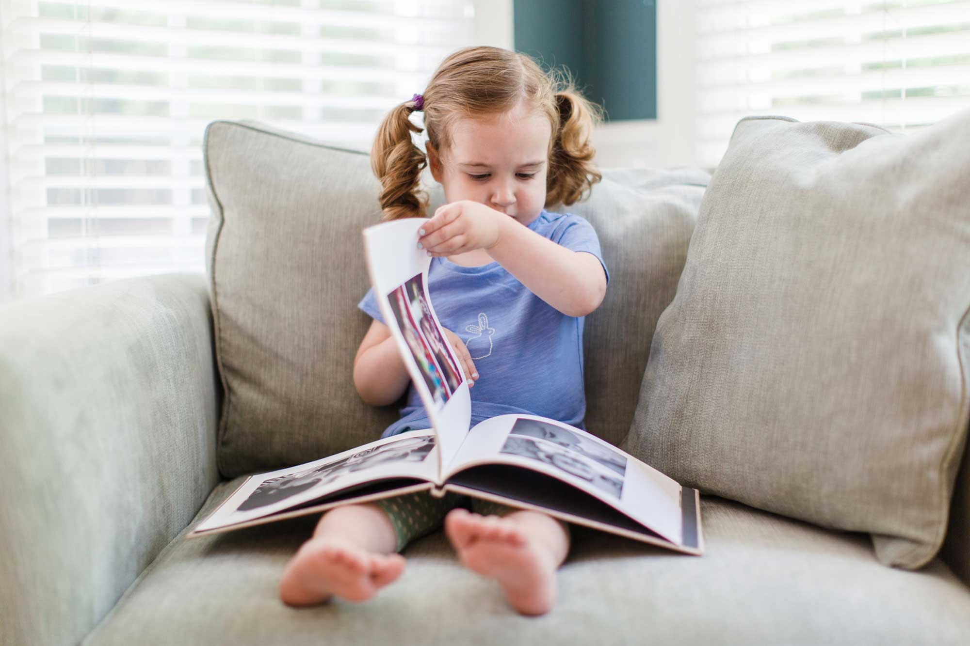 Young girl turning the pages of a printed hardcover photo book on a sofa